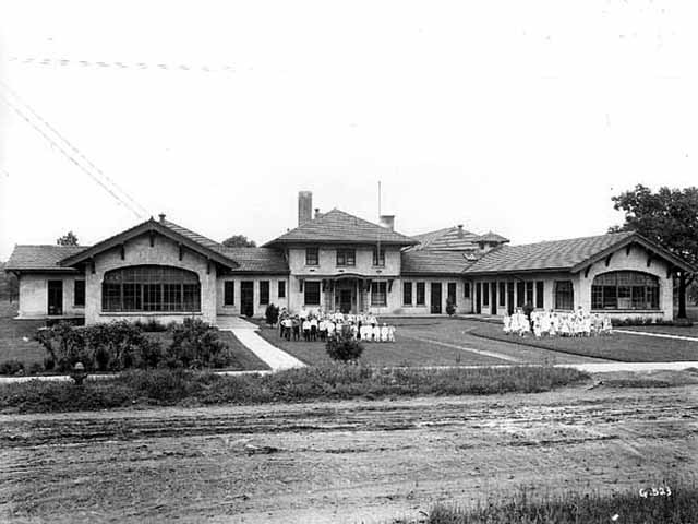 Black and white photo shows a beautiful building with several wings and a spacious lawn. Two groups of children are on the lawn, with boys to the left and girls to the right. Both groups appear to be wearing uniforms.
